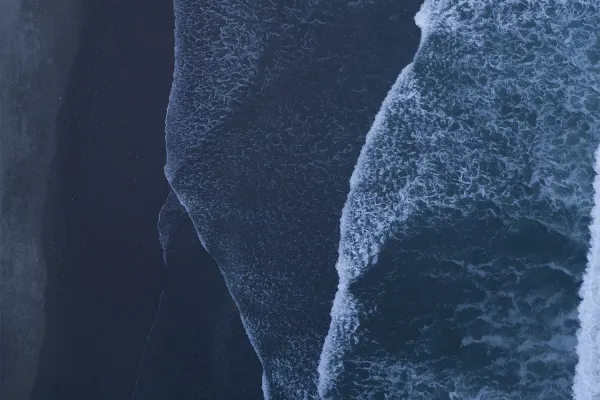 Aerial view of dark ocean waves crashing onto a black sand beach, with a small boat in the lower left corner.